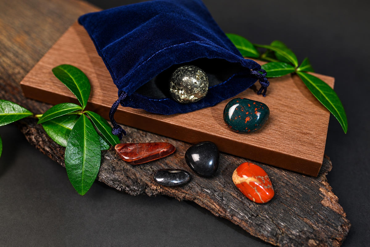A blue velvet pouch on a wooden board holds a metallic sphere. Five polished stones in red, black, green, and orange sit on wood and bark with green leaves.