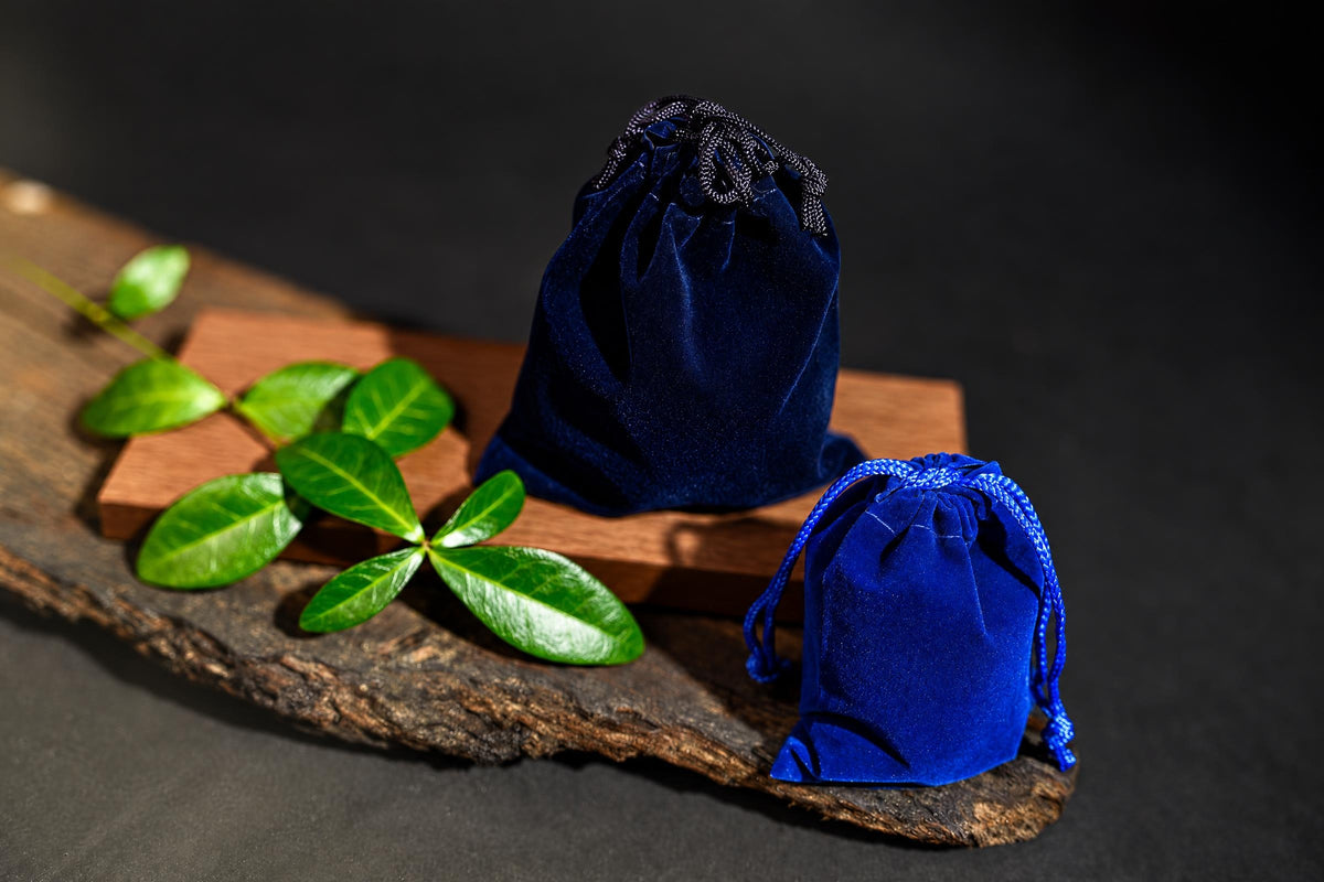 Two blue velvet drawstring pouches sit on a wooden surface with green leaves nearby.
