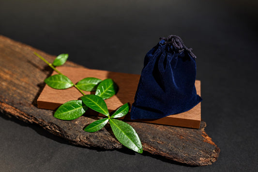 A dark blue velvet pouch with a drawstring sits on a wooden block, next to green leaves on a rustic wood slab.