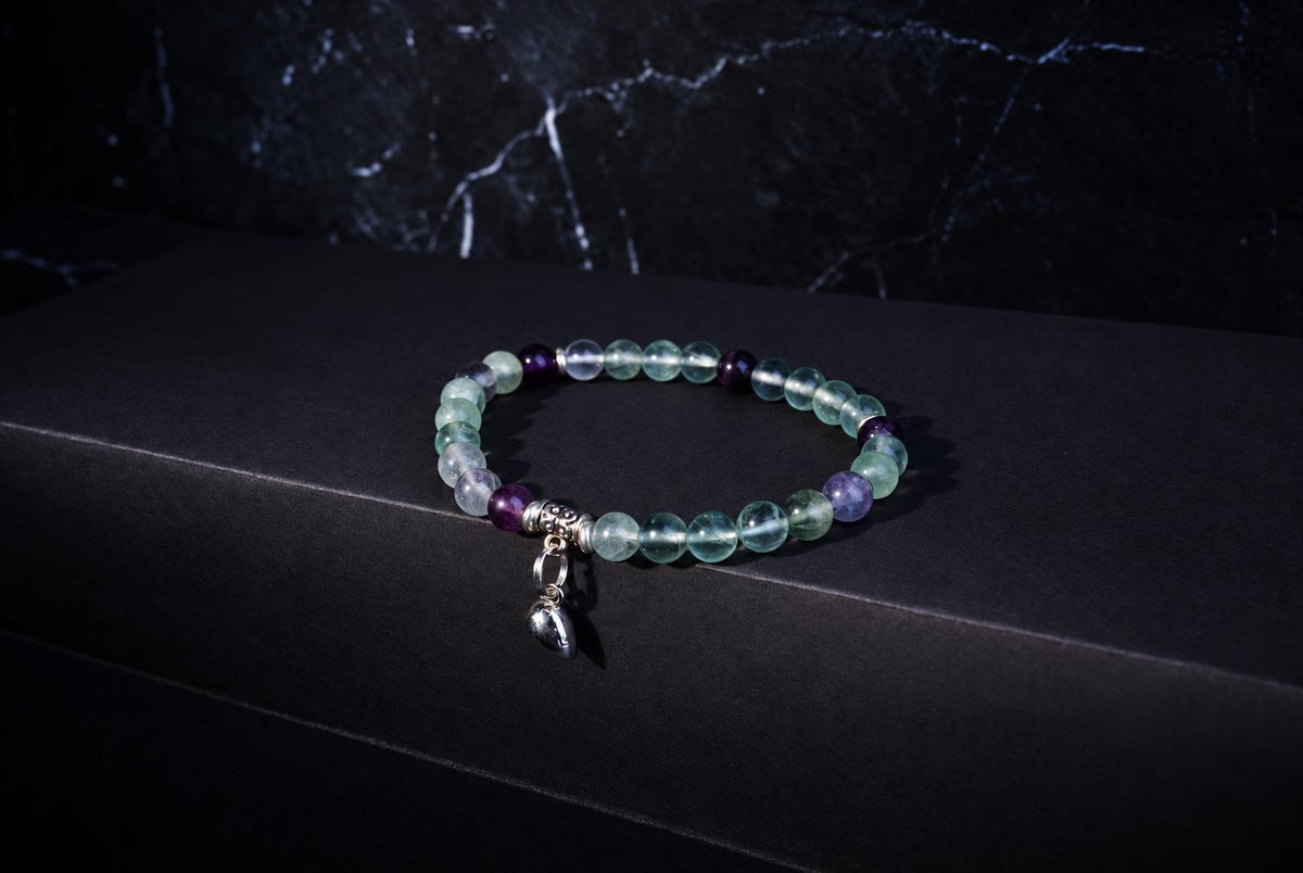 Fluorite bracelet with translucent green and purple round beads, ornate silver spacers and a hanging heart charm on a dark background.