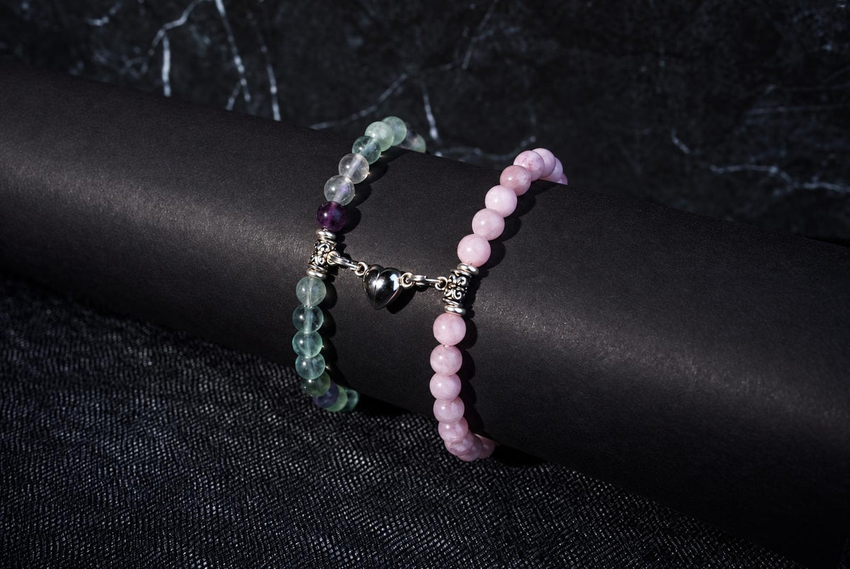 Two beaded bracelets on a dark cylindrical form: one with green and purple beads, the other pink, joined by a silver heart charm.