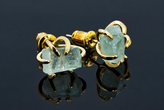 Two gold-coloured stud earrings with pale blue-green rough fluorite stones held by curved metal claws on a dark reflective surface.