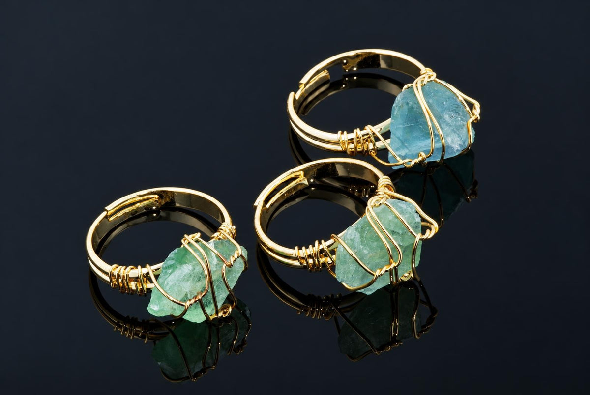 Gold-coloured rings hold rough green and blue crystals wrapped in thin wire on a dark reflective surface.