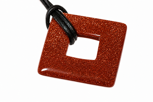 A square, donut-shaped pendant with a glittery orange-brown surface and a central hole, attached to a black cord against a white background.