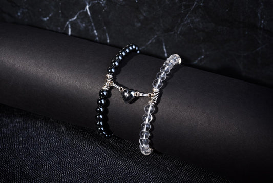 Two bracelets on a dark surface: one with black beads and a silver heart charm, the other with clear beads.