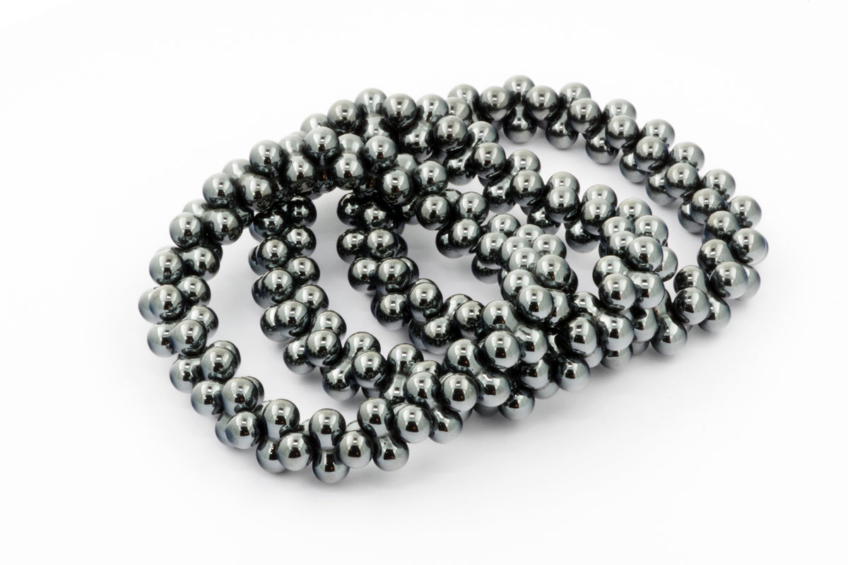 Close-up of multiple strands of polished dark gray hematite beads arranged loosely on a white surface, showcasing the natural crystal’s reflective and smooth texture, Hematite bracelet – DNA.