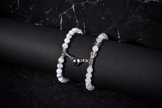 Two bracelets on a dark cylindrical form: one with white marbled beads, the other with clear faceted beads; both have silver spacers and a shared heart-shaped charm.