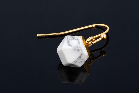 A gold-coloured hook earring with a white, grey-veined faceted cube stone on a dark reflective surface.