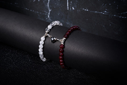 Two beaded bracelets, one clear and one red, with a silver heart charm, rest on a dark cylindrical surface.