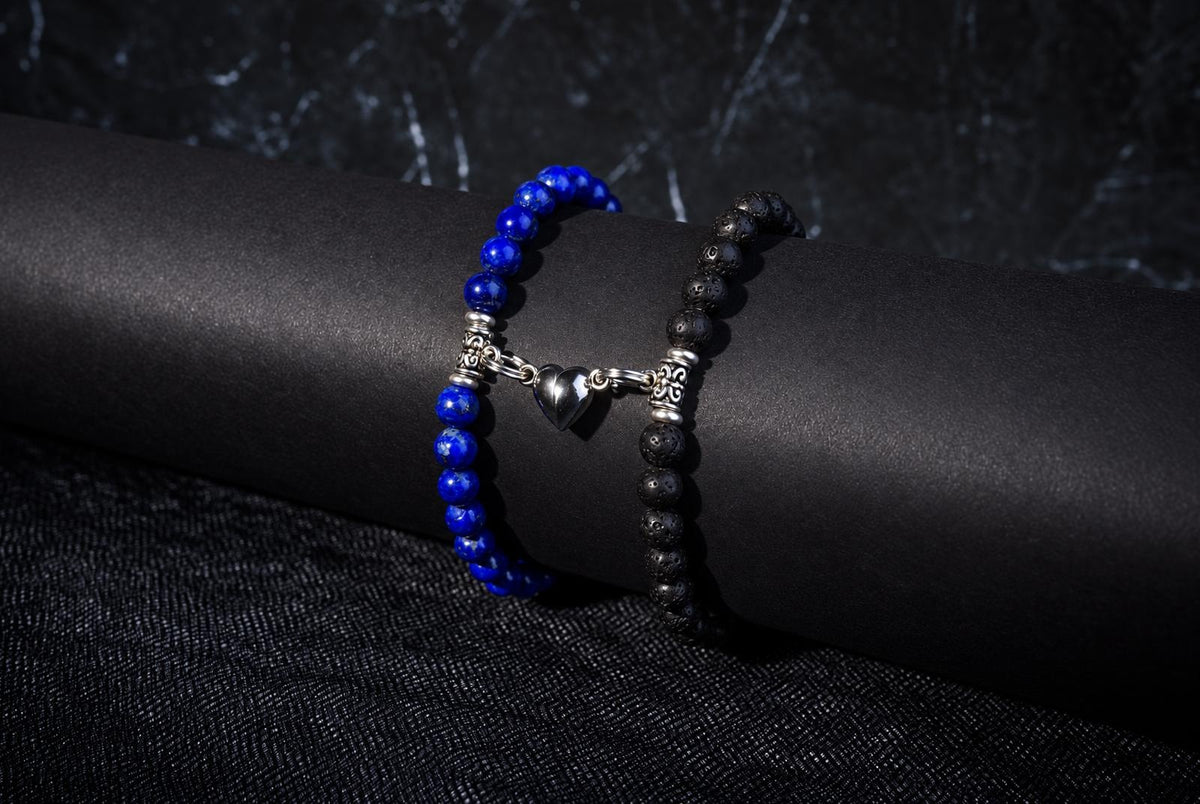 Two beaded bracelets on a dark surface: one with blue beads and silver heart charm, the other with black porous beads.
