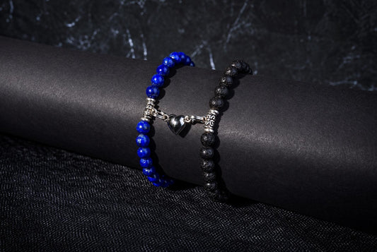 Two beaded bracelets on a dark cylindrical form: one with blue beads, the other black, joined by a silver heart charm.
