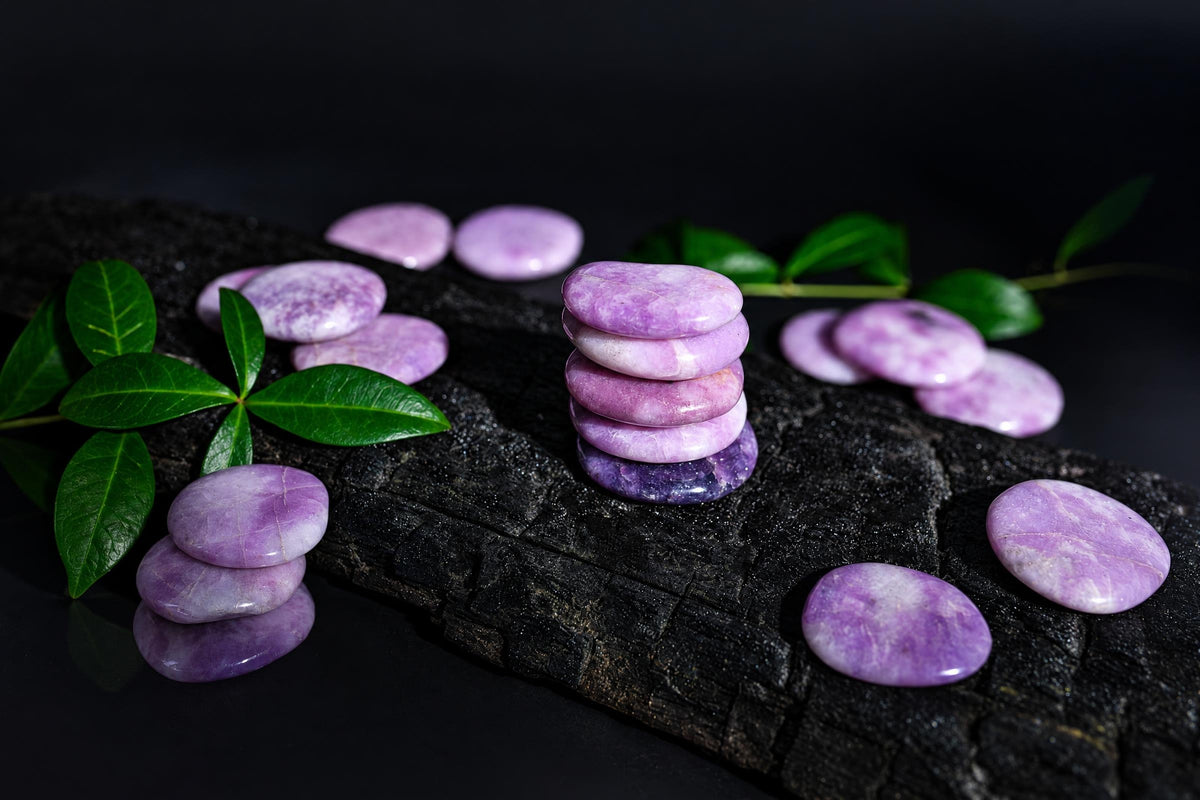 Polished purple oval stones are stacked and scattered on a dark textured surface among green leaves.