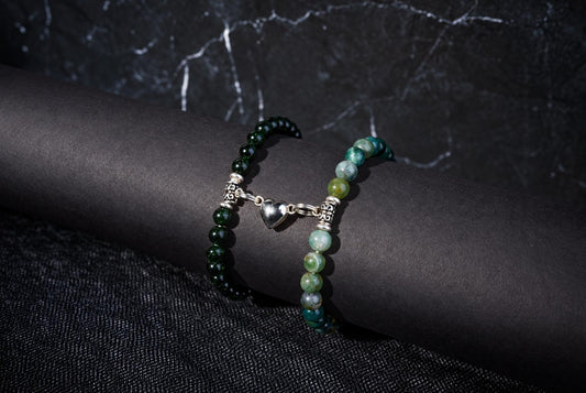 Two beaded bracelets with dark and pale green stones, ornate silver spacers and linked heart charms on a dark background.