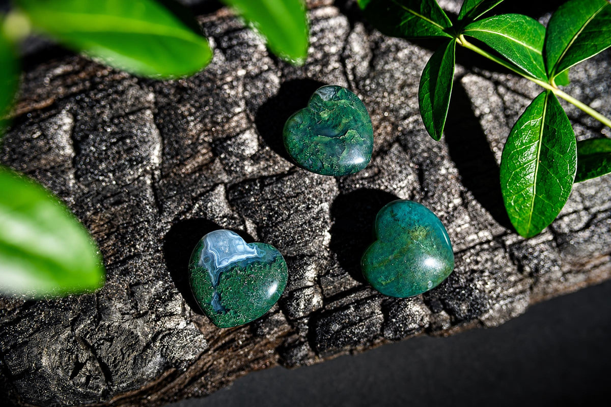 Polished green heart-shaped stones with pale patterned patches rest on a dark textured surface among green leaves.