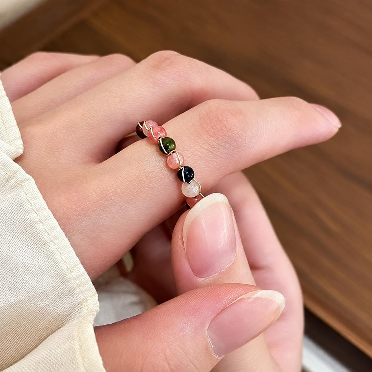A hand wears a thin, open ring with multiple round beads in pink, green, black and white on a wooden surface.