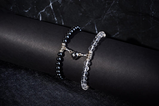 Two beaded bracelets resting on a black cylindrical display stand. One bracelet features opaque black beads and the other features clear, transparent beads. Both are connected by a silver metal charm shaped like a heart.