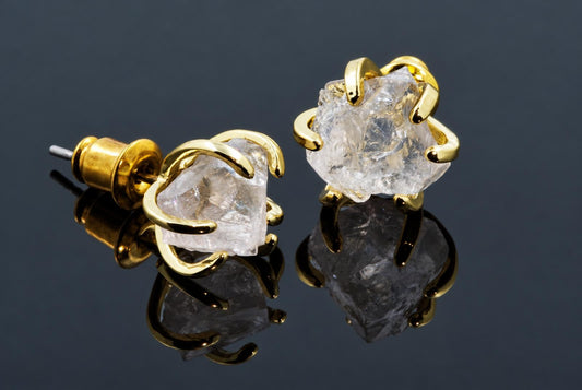 A pair of gold-coloured stud earrings with claw-set, rough quartz stones on a dark reflective surface.