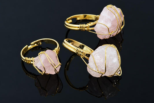 Gold-coloured rings hold rough pale pink crystals wrapped in thin wire on a dark reflective surface.