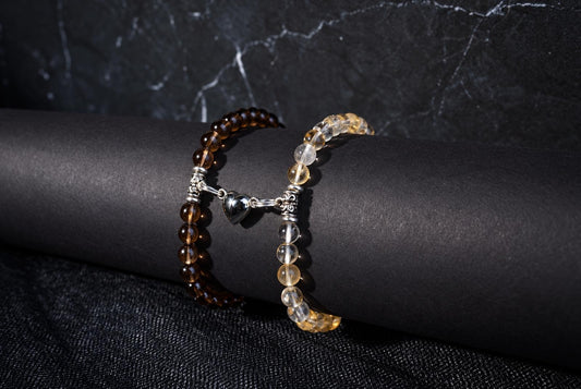 Two bracelets of dark brown and clear yellow beads joined by a silver-tone heart charm on a black display.