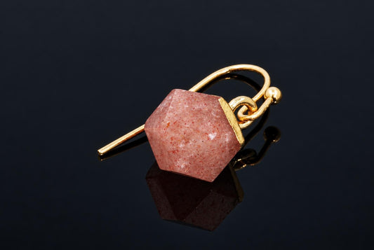 A pink speckled faceted cube earring with a gold-coloured cap and hook rests on a dark reflective surface.