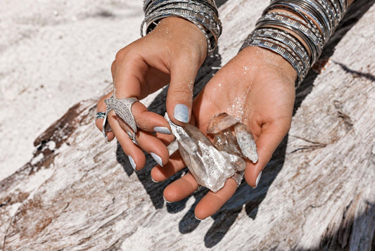 Hands wearing metallic rings and bangles hold clear rough crystals above a pale wooden surface.