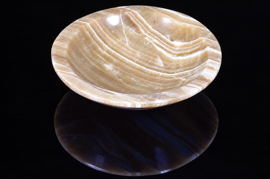 A polished, oval bowl with wavy beige and white stripes sits on a dark, reflective surface.