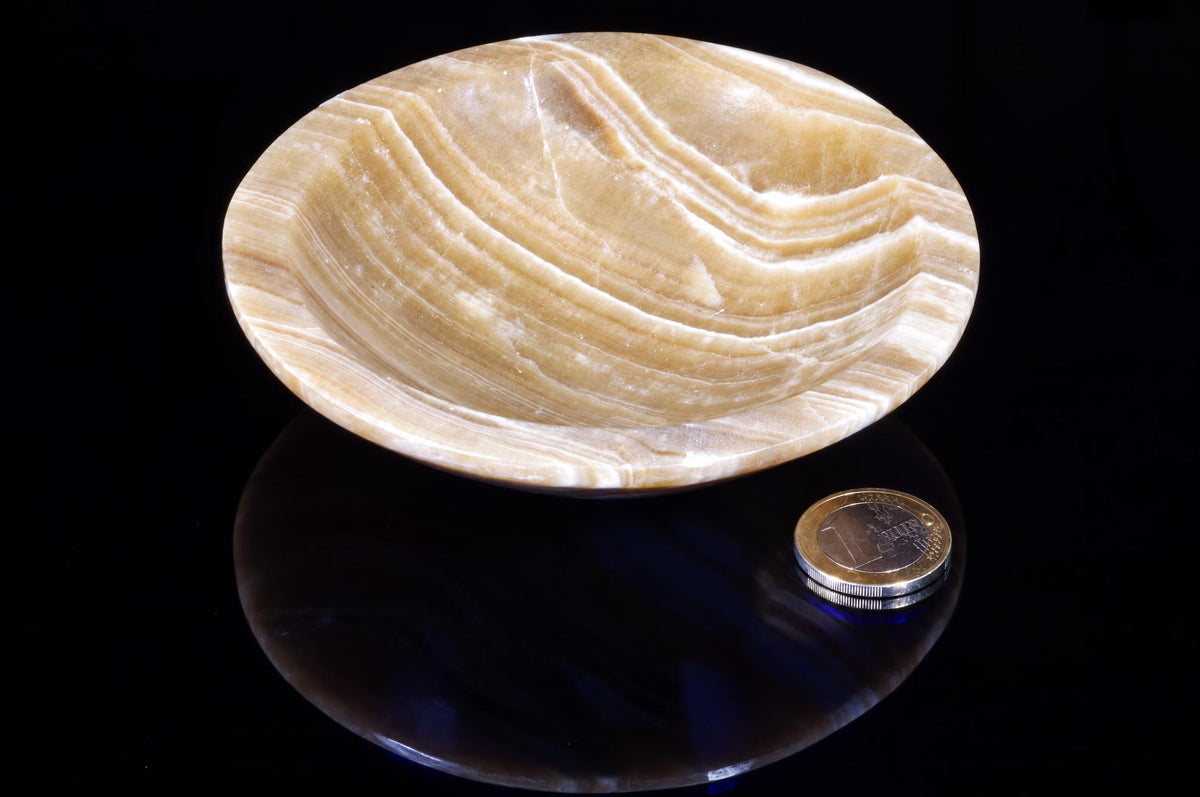 A polished, oval bowl made of layered beige and white stone sits on a dark surface next to a coin.