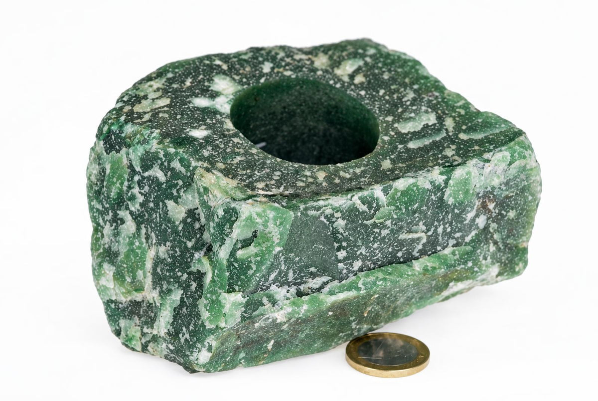 A green and white speckled stone object with a central hole, resting on a pale surface next to a coin.