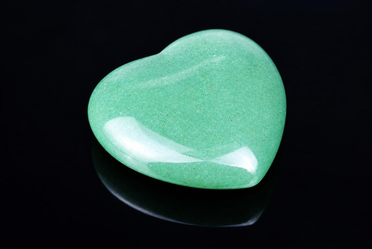 A polished, heart-shaped green aventurine stone with a smooth surface and subtle sparkle rests on a dark reflective surface.