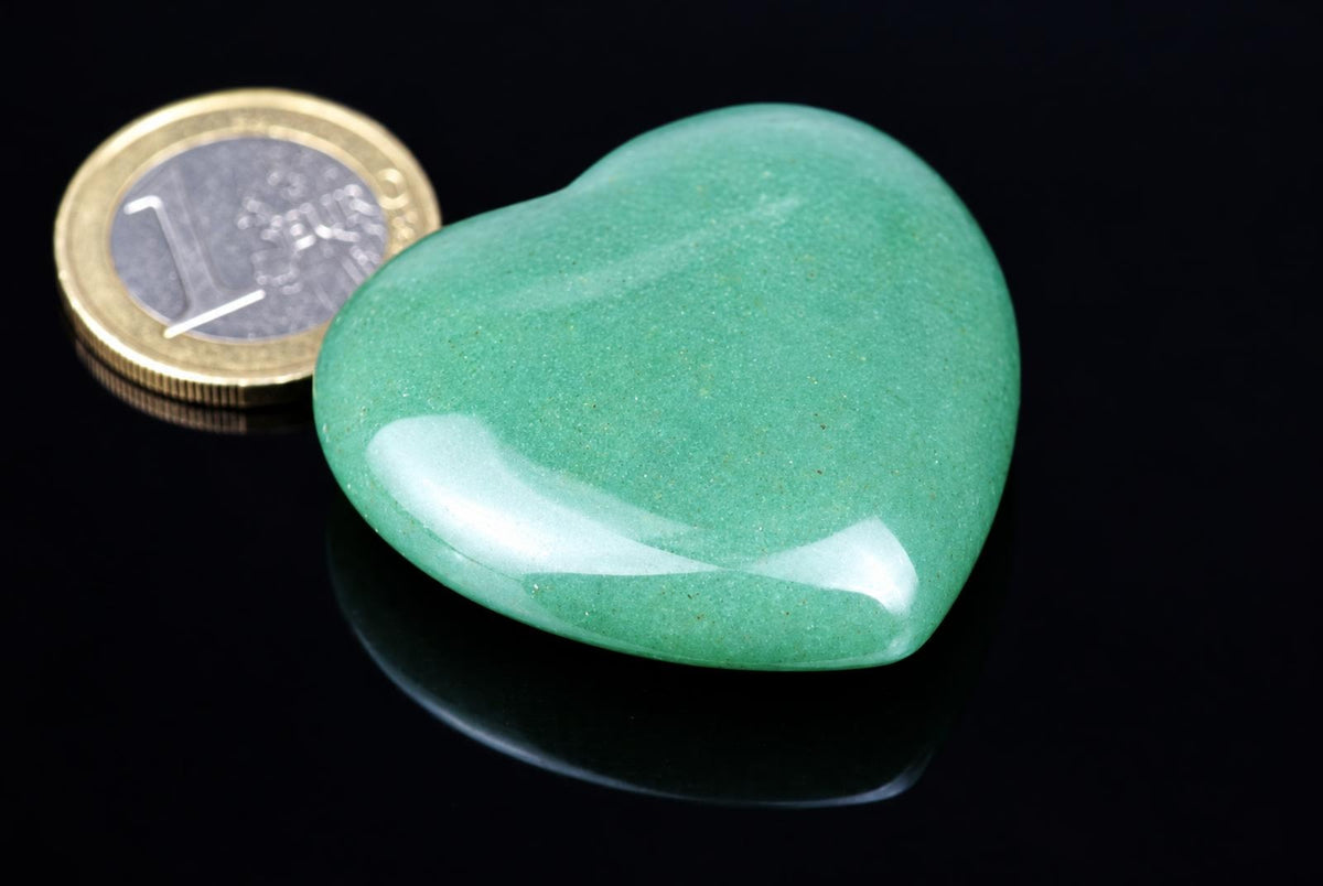A polished green heart-shaped stone sits on a dark surface next to a one-euro coin for scale.