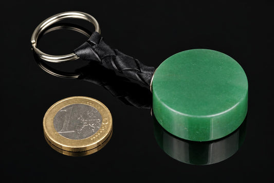 A green aventurine keychain with a braided black leather strap and metal ring, shown on a dark reflective surface next to a one-euro coin.
