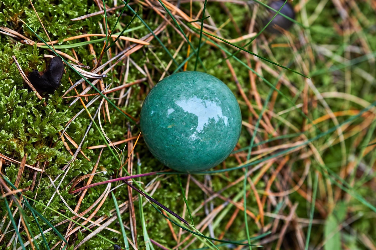 A polished green aventurine sphere rests among moss, grass and thin brown stems.