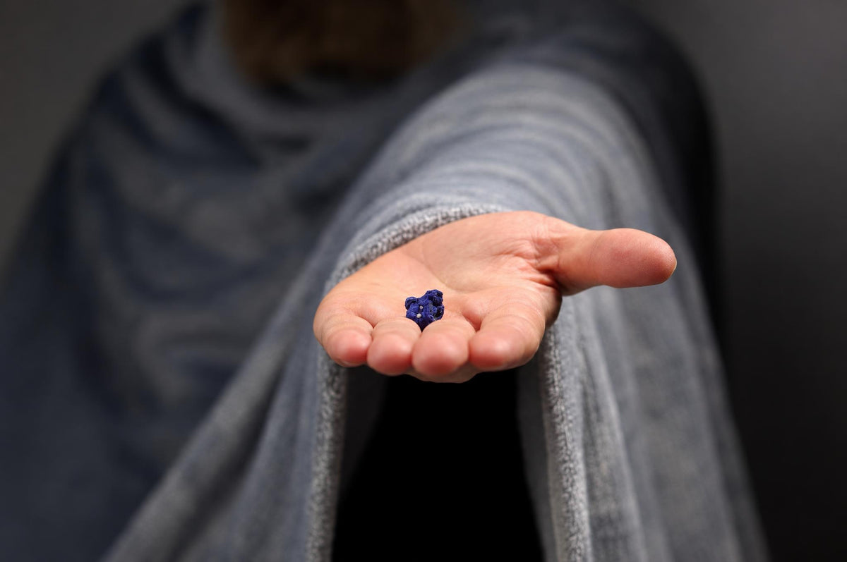 A person in a grey robe holds a small, dark blue Azurite crystal with white flecks in their open palm against a dark background.