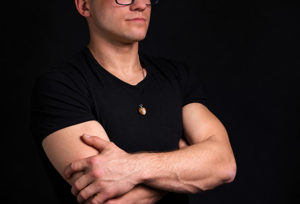 A man in a black t-shirt wears a necklace with a small, oval-shaped pendant featuring brown and cream bands against a dark background.