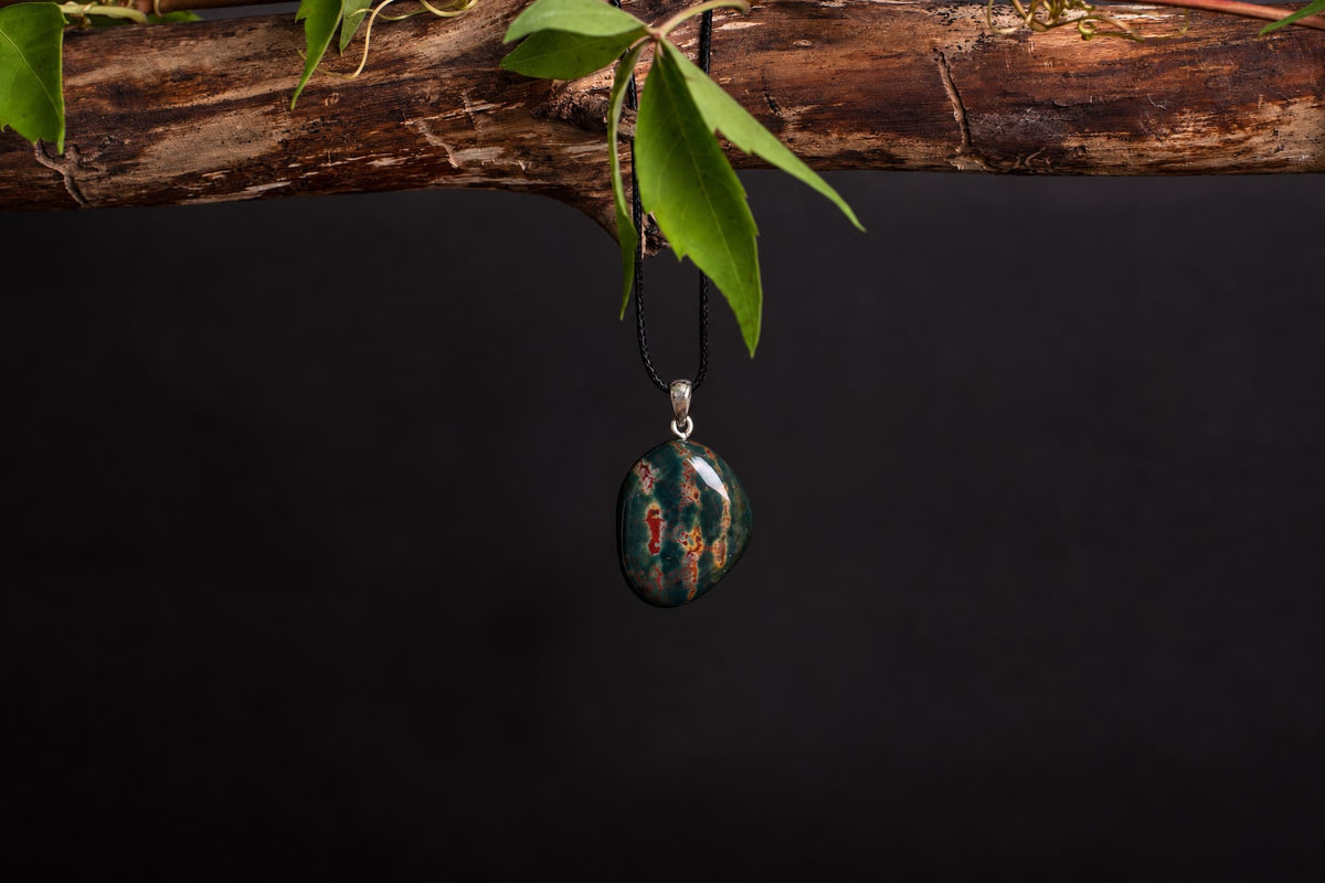 A polished oval pendant with green and red patterns hangs from a black cord on a dark surface.