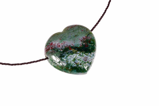 A heart-shaped pendant with green, red and white speckled stone on a dark cord against a pale surface.
