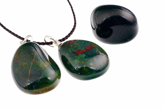Three polished green stones with red speckles, two on dark cords and one loose, against a white background.