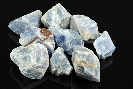 A pile of raw blue calcite crystals with white and brown inclusions on a dark reflective surface.