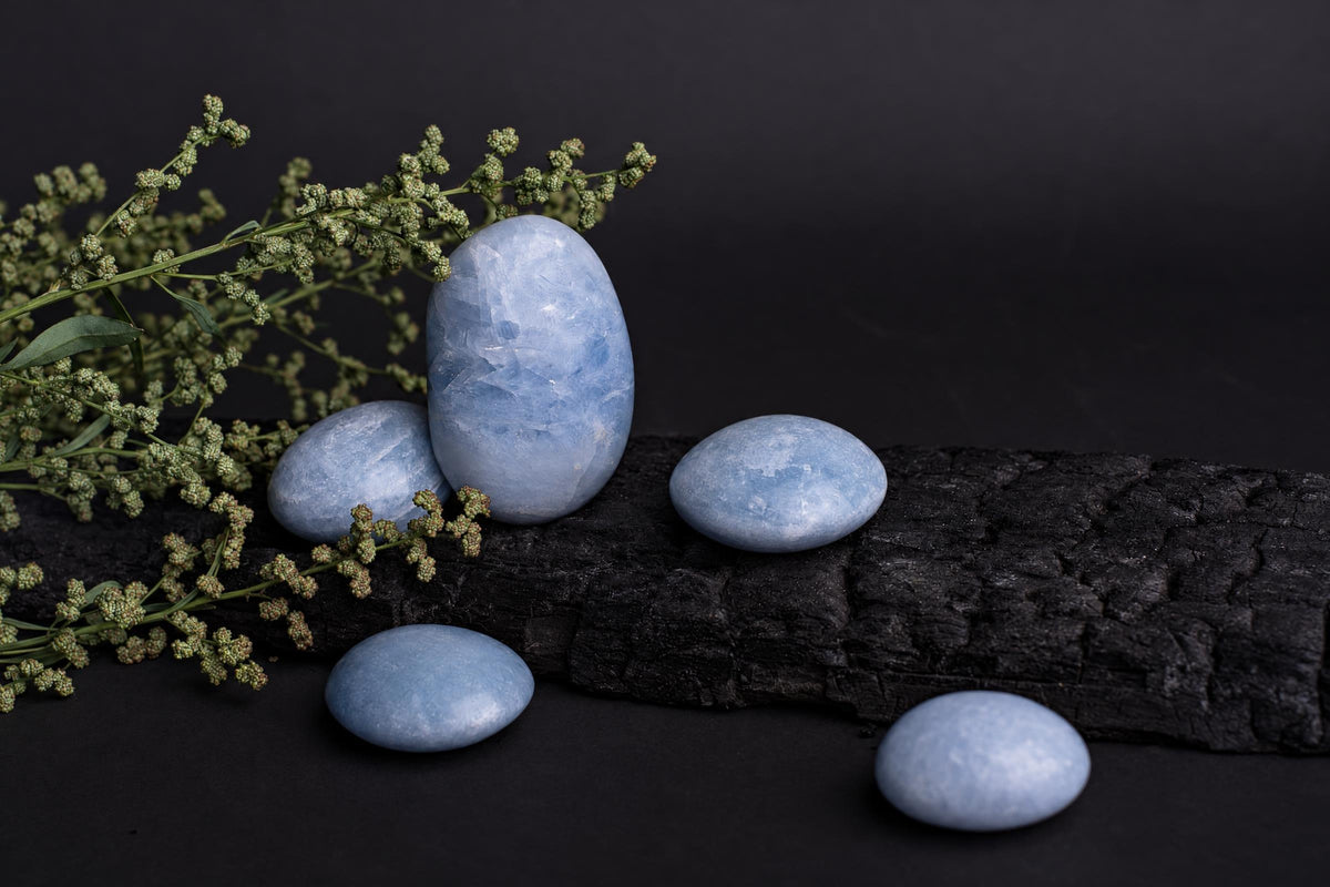 Five smooth, pale blue stones rest on a dark wooden surface beside green sprigs against a black background.