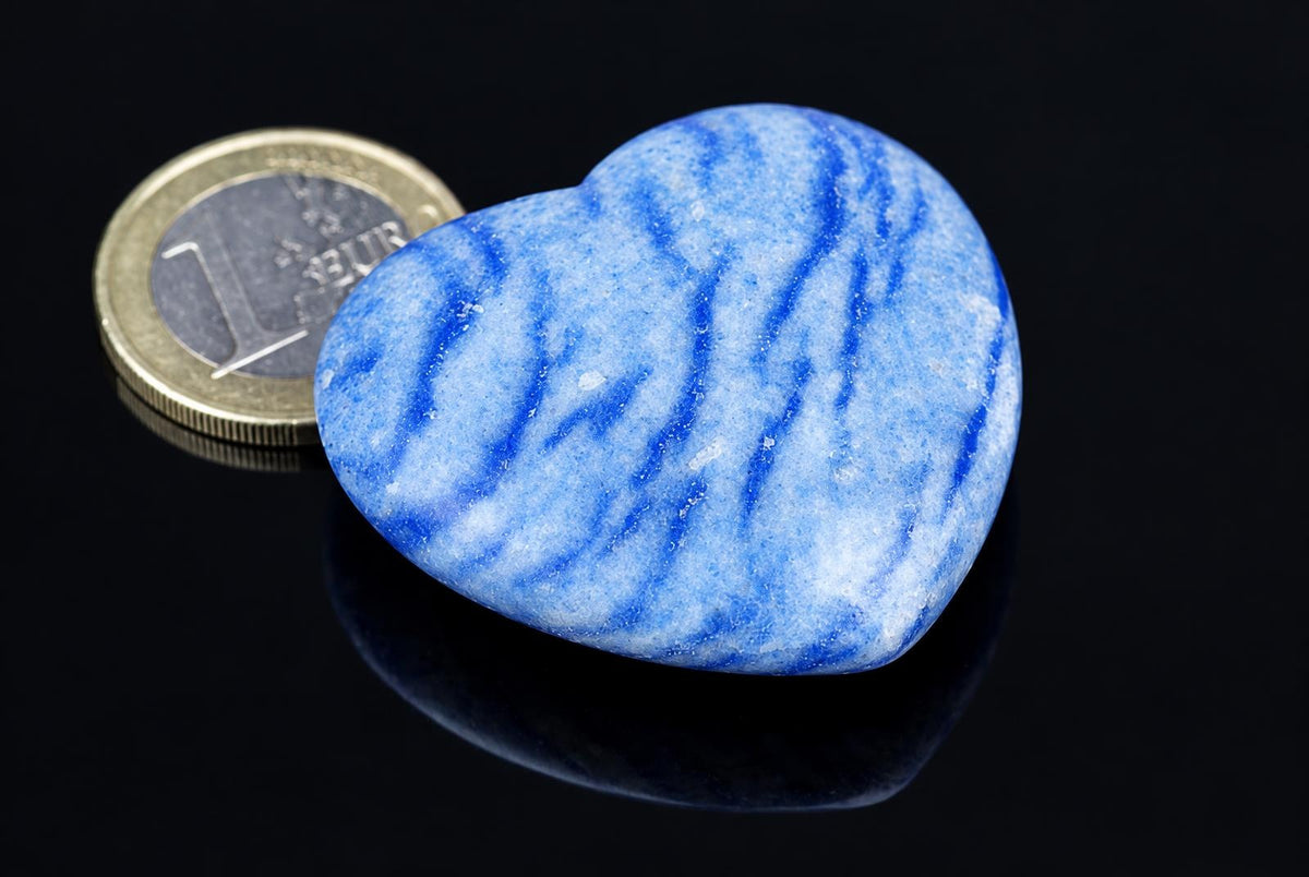 A polished blue quartz heart with white streaks rests on a dark surface next to a one-euro coin.