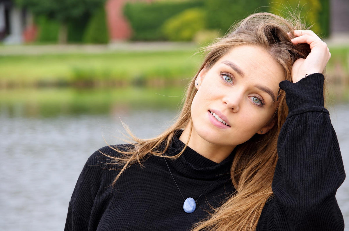 A woman wears a black turtleneck sweater with a blue quartz pendant on a thin cord, outdoors near water.