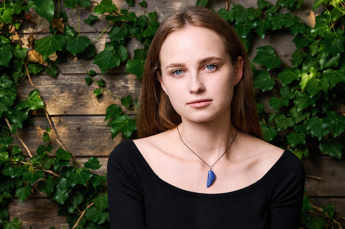 A woman wears a black top and a necklace with a blue pendant shaped like a wing, set against a wooden wall with green ivy.