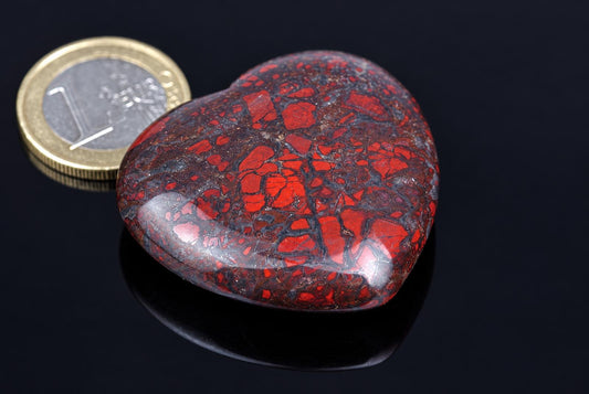 A polished heart-shaped stone with red and dark brown brecciated patterns rests on a dark surface next to a coin.