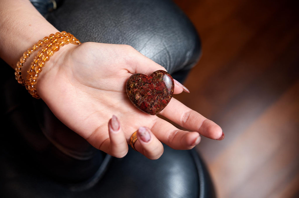 A hand holds a polished, heart-shaped stone with red and brown speckled patterns on a dark surface.