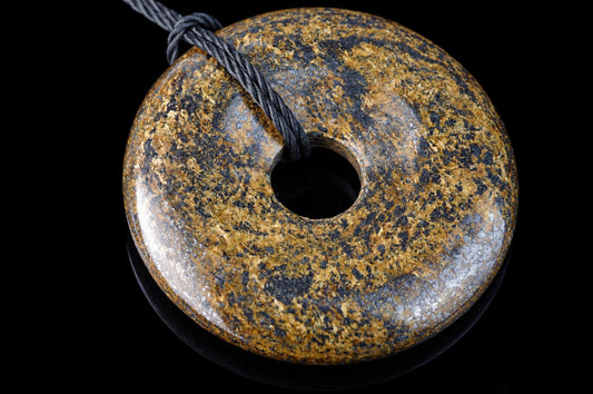 A polished, donut-shaped pendant with a speckled brown and black surface, featuring a central hole and a dark cord looped through it against a black background.