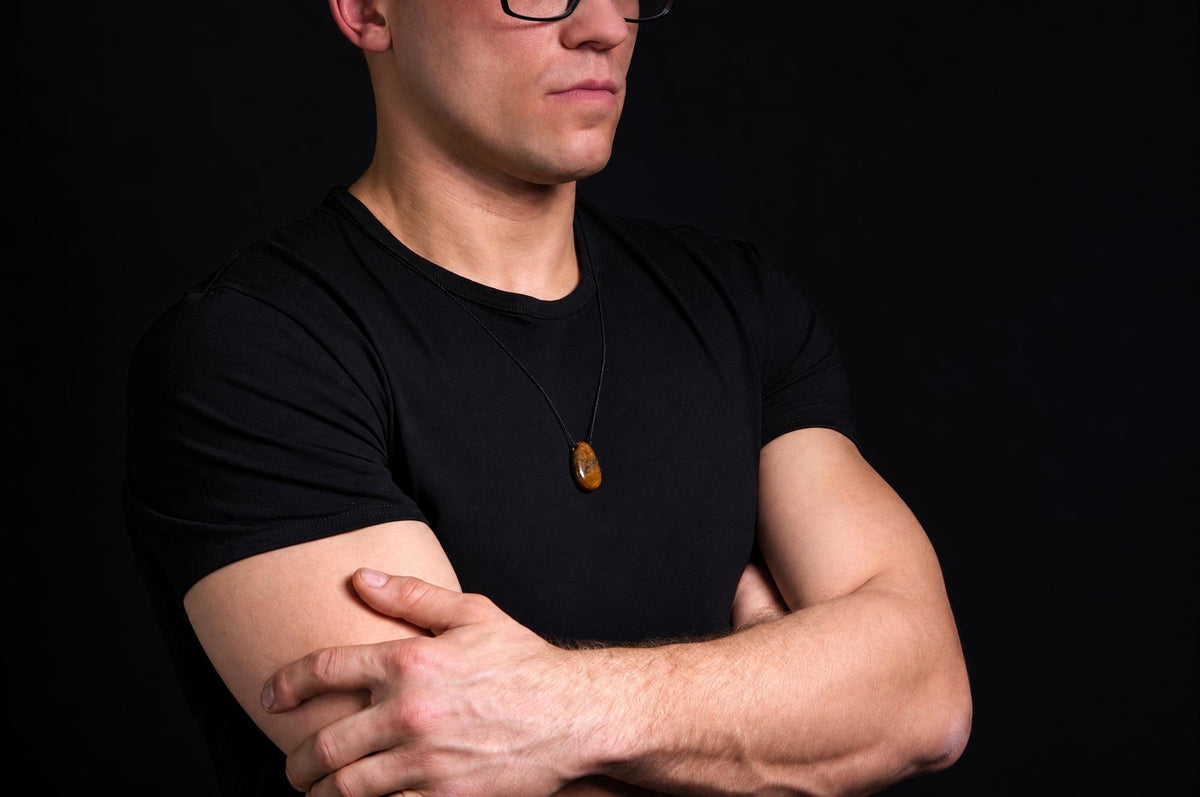 A man in a black t-shirt wears a necklace with an oval, brownish pendant against a dark background.