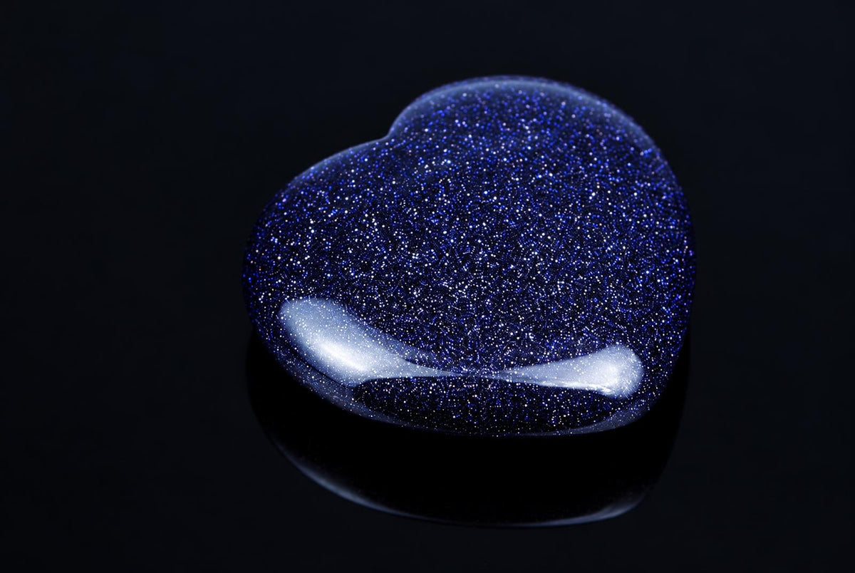 A heart-shaped object with a dark blue, glittery surface and white highlights rests on a dark, reflective surface.