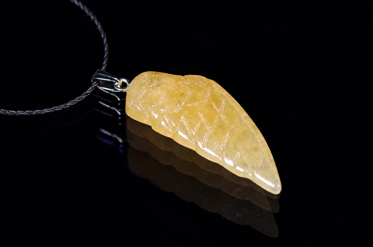 A yellow calcite pendant shaped like a wing, carved with lines, on a dark surface.