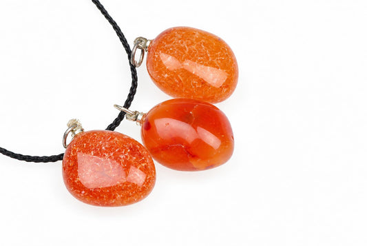 Glossy orange carnelian pebble pendants with translucent interiors lie together, with one attached to a black cord against a white background.