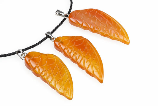Orange carnelian wing pendants with carved feather lines are arranged with silver toned loops and black cords against a white background.